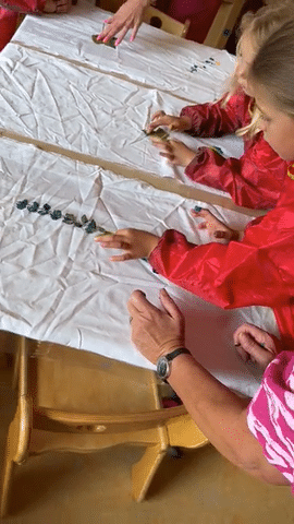 Kinder und eine Erzieherin drucken bemalte Blätter auf weißen Stoff