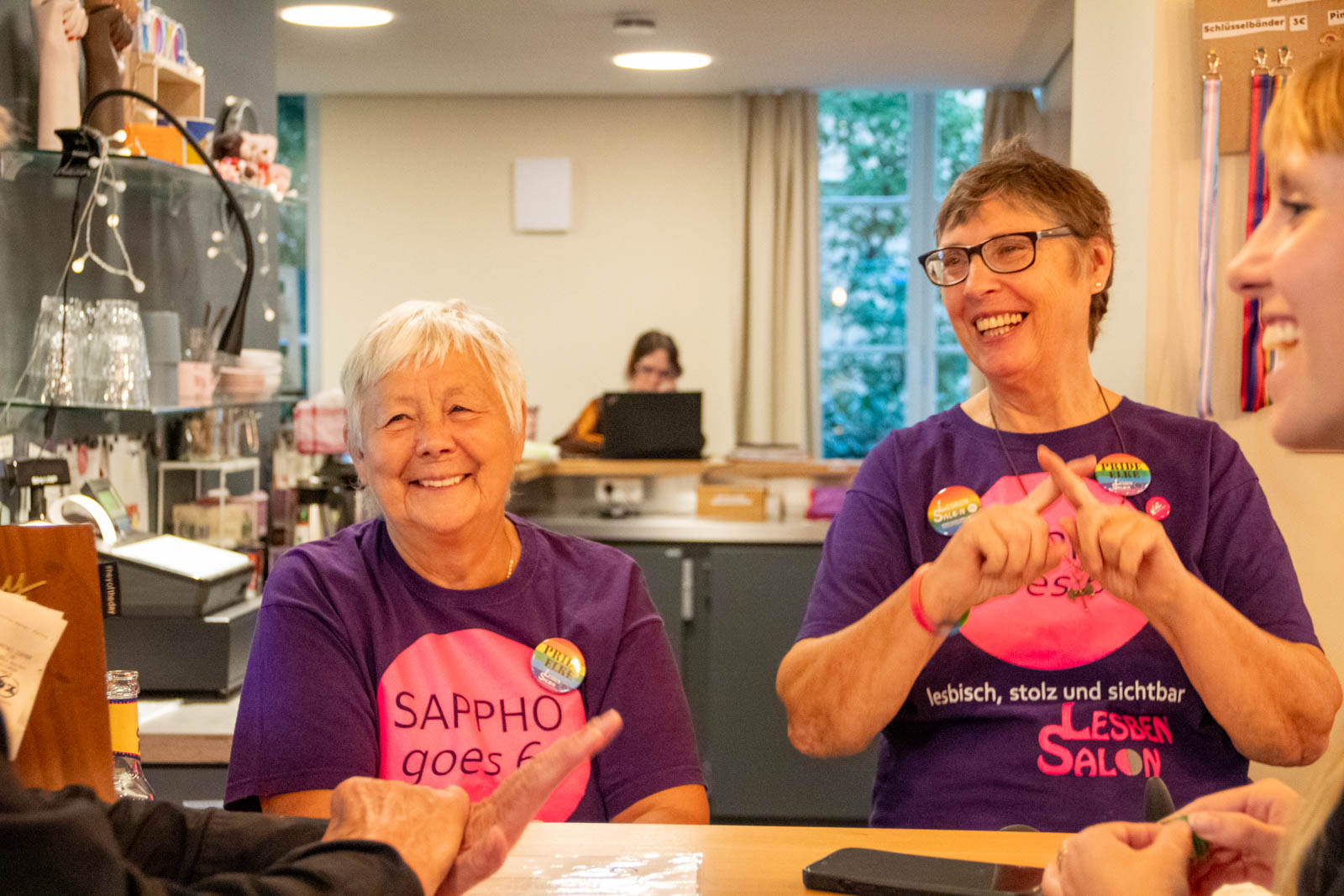 Zwei Frauen in Sappho-goes-60-T-Shirts lachen am Tresen