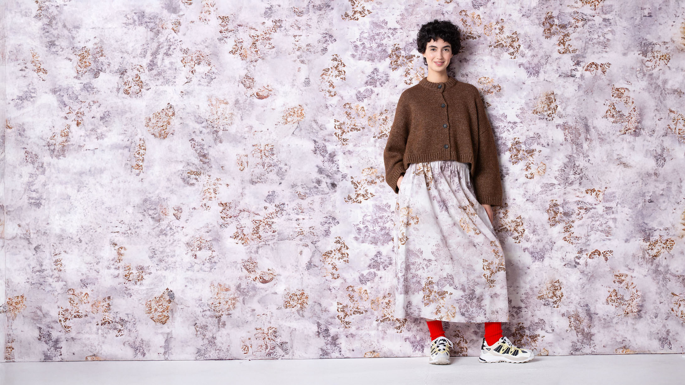 Frau in brauner Strickjacke und geblümtem Rock vor einer Blumentapete