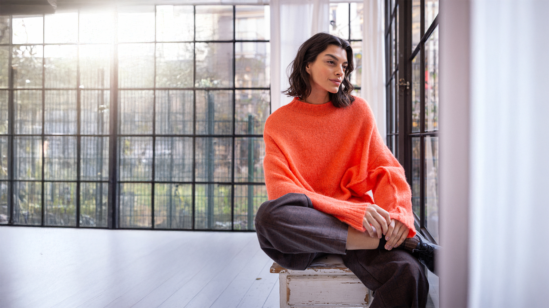 Frau in orangem Strickpullover und weiter Hose sitzt auf einem Hocker vor einer großen Fensterfront